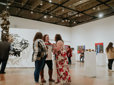 A group of people gather and talk in a brightly lit art gallery featuring abstract paintings and sculptures on pedestals.