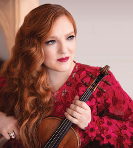 Rachel Barton Pine sits for her professional photo, wearing a reddish-pink dress and holding her violin.