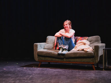 A USD student sits curled up on a worn gray sofa on a dark theater stage, smiling softly beside a folded blanket.