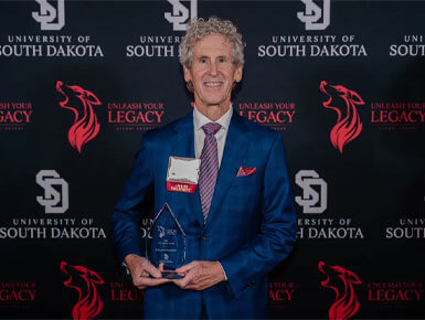Pierce H. McDowell holds his alumni award at the USD Unleash Your Legacy Awards ceremony, standing before a “Leaving Your Legacy” backdrop. He is wearing a blue suit.