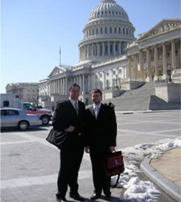 Logan Hollers and a friend stand, who are both wearing professional suits, stand in front of the U.S. Capitol building on a sunny day.