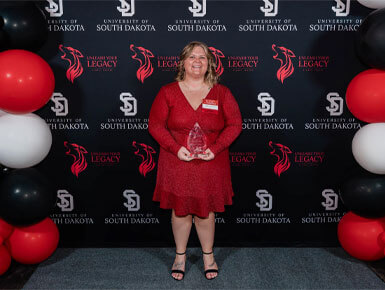 Rachelle Norberg smiles in her red dress as she holds an award at the USD Unleash Your Legacy Awards ceremony. She is posed before a USD branded backdrop framed by red, white and black balloons.