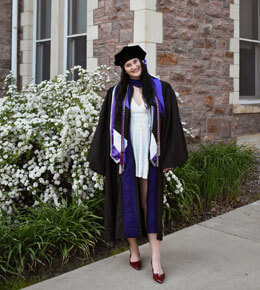 Kylee VanEgdom stands in front of Old Main on the USD campus, wearing her graduation regalia. She is smiling at the camera.