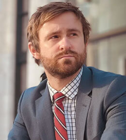 Logan Hollers poses for his professional photo, sporting a beard and short brown hair, wearing a grey suit jacket, a checkered white and blue shirt, and a red and white striped tie. He is looking off into the distance.