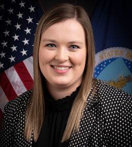 Kelcy Schaunam poses for her professional photo, wearing a black and white polka dot blazer, standing in front of an American flag and a government department flag.