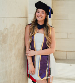 Mayson Coyle stands against a pillar in her graduation cap, sash and cords. She is smiling at the camera.