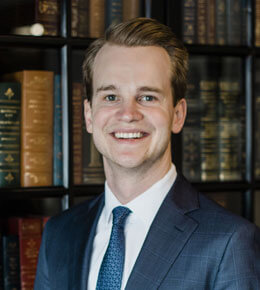 Pete Heidepriem stands in front of a bookshelf, smiling at the camera for his professional photo. He is wearing a blue suit and tie.
