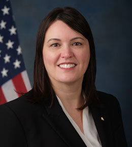 Professional photo of Jackie Barber with dark hair, smiling and wearing a black blazer and white top, standing in front of a blurred American flag backdrop.