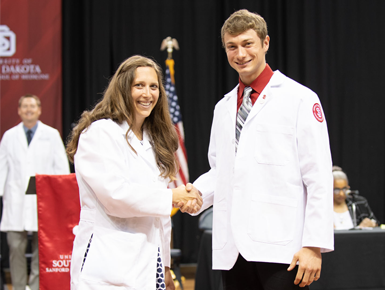 Two people wearing medical white coats shake hands and smile toward the camera.