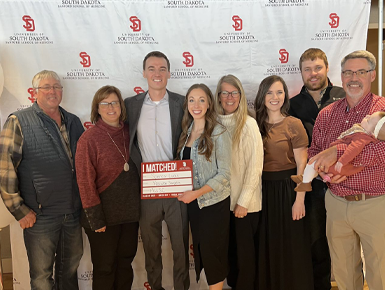 A medical student stands with his family and smiles for a photo. The student is holding a marker board that shows which residency program they matched into. 
