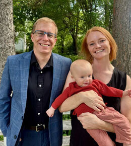 Jonathon Gruba stands in a blue blazer and glasses, with his partner, and a baby in a red sweater smiling at the camera, standing outdoors near wooden structures.