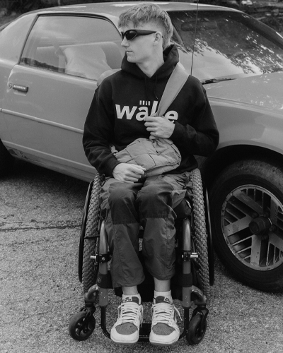 A person in a wheelchair in front of a car. They wear sunglasses and look off to the side.