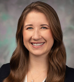 Ellie Blue smiles at the camera for her professional photo. She is wearing a necklace and a black blazer with a white tank top underneath.