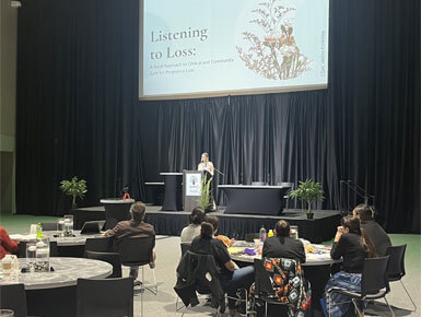 Rebecca Hofer stands on a stage, in front of a podium, giving a presentation. Behind her is a screen that reads "Listening to Loss."