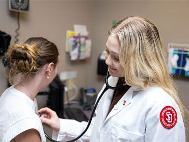 A USD SSOM student listens to a patient's heartbeat with her stethoscope in a clinic room.