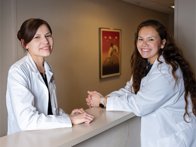 Drs. Arna Mora and Carol Whitman stand facing each other while leaning their arms on a counter. They are both in white coats.