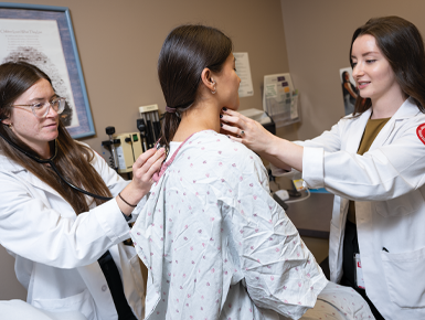 Two medical students in white coats examine a seated patient. One student checks the patients next, while the other holds a stethoscope to the patient's upper back. 