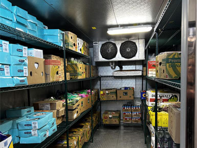This photo shows the inside of a cooler or walk-in freezer, with shelfs lined with canned food, produce, and more.