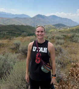 AJ Uthe smiling in a black "UTAH 6" tank top and fanny pack, standing in a grassy mountain landscape under a bright sky.