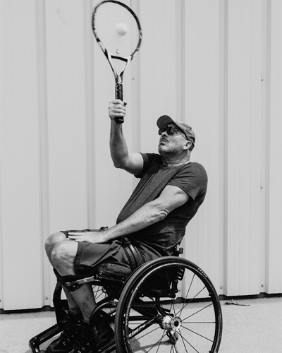 A person in a wheelchair holds up a tennis racket and hits a ball.