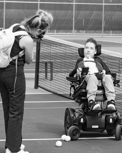 A person who is standing takes a picture of a person who is in a wheelchair on a tennis court in front of the net.