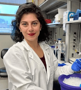 Dela Moradpour stands in a laboratory, with a white lab coat on and blue gloves, with dark curly hair and red lipstick, smiling.