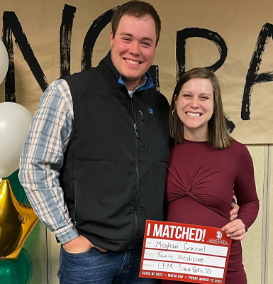 A medical student stands with her husband and smiles for a photo while holding a marker board that shows which residency she matched with.