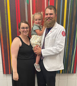 USD SSOM student Aaron Fest stands in a white lab coat, with his partner in a black dress, and a small child standing together against a background of vertical colored stripes.