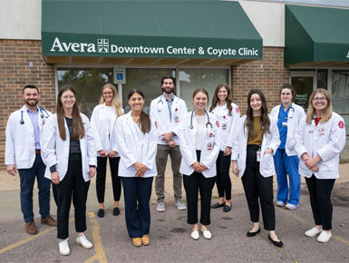 USD SSOM students stand in front of the Coyote Clinic. They are wearing their white coat attire, with stethoscopes.