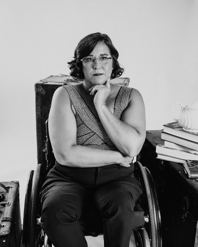 A person in a wheelchair has their hand under their chin in question and looks toward the camera. There is a stack of books on a table on the right edge of the photo.