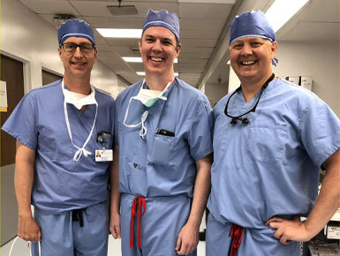 Dr. John Lee, far right, stands with Dr. Doug Trask and Dr. Eric Wilkinson. They are dressed in blue surgical scrubs and hats.