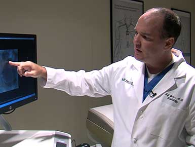 Dr. AJ Gunn wears a white lab coat and points at a screen in a medical office.