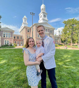 Parker Owen stands, with his family, in a white lab coat on a grassy lawn in front of Old Main.
