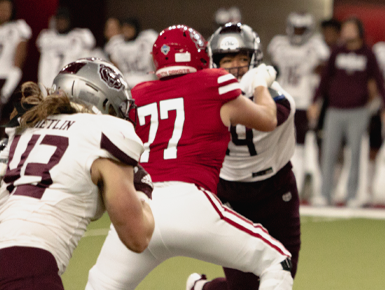 A football player wearing a red jersey with the No. 77 blocks a player wearing a white jersey.