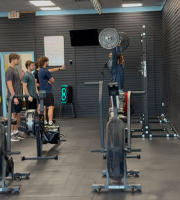 In a fitness gym, a student exercises on a strength machine while several people watch beside the weights, creating a focused training atmosphere.