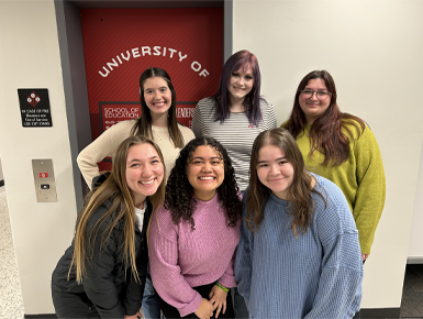 Six USD students from the School of Education stand together in front of a red University of South Dakota School of Education door, smiling at the camera.