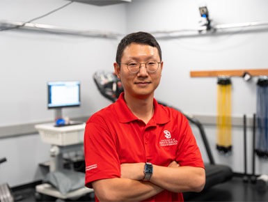 Hyung Suk Yang stands posing for a photo. He is wearing  glasses, a red USD polo shirt and standing with his arms crossed. He is in a sports science laboratory. Behind him, a treadmill, a metabolic cart with a computer screen, and resistance bands are visible, indicating a research-focused environment.