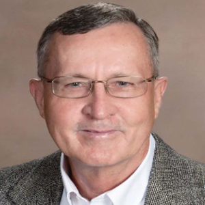 Ted Williams, Ed.D. ’84, looks at the camera smiling for his photo, with a light brown background.