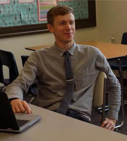 Sean Binder sits in a classroom with a laptop sitting in front of him. He is smiling and looking off into the distance.