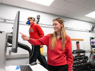 Sydney Conley stands in the KSM lab. She is wearing a red USD sweatshirt and pointing to a computer monitor. In the background, a male subject wearing a blue metabolic mask and red sweatshirt walks on a treadmill. The room is equipped with various fitness equipment like dumbbells and medicine balls.