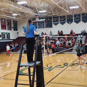A volleyball referee stands on the referee stand next to the net, watching the match in progress.