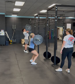 A student performs a barbell squat in a fitness gym, while another student observes, standing beside squat racks and weight plates. 
