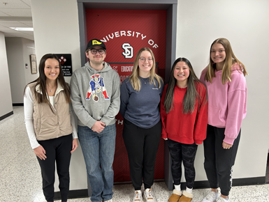 Five student teachers from the USD School of Education stand in front of a red USD door. They are smiling and facing the camera.