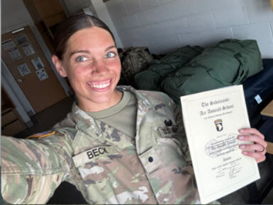 Danielle Beck poses for a selfie, holding a certificate type document in her left hand. She is standing in a dorm-like room, smiling at the camera.