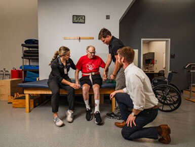 An older gentleman sits on a patient bed with a band around his waist. He is surrounded by physical therapists, who are helping him. 