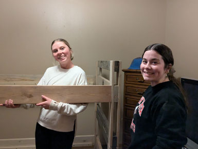Two USD students smile at the camera while working on a woodworking project indoors. One student holds a long, unfinished wooden plank against a vertical wooden frame, as part of the project they are assembling.