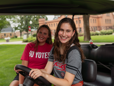 Two USD students sit in a Charlie Cart and smile for a photo on campus.