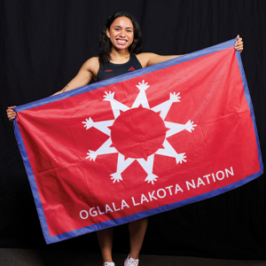 Matayah Yellow Mule poses with the Oglala Lakota Nation Flag, which is red and white, with a blue outline.