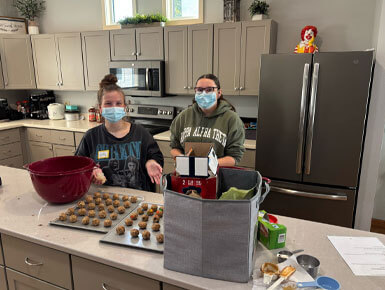 Two USD students, who are wearing light blue surgical masks, stand behind a kitchen island at a Ronald McDonald House. They are preparing food; on the counter, there are two baking sheets filled with neatly rolled dough balls and a large mixing bowl. A small Ronald McDonald figurine is visible sitting on top of the stainless steel refrigerator behind them.