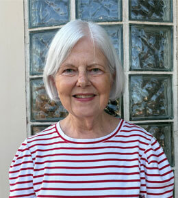Susan Tuve stands in front of a window for her headshot photo. She is wearing a red and white shirt.